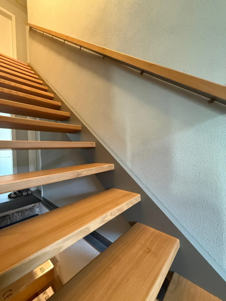 Eine moderne Holztreppe mit offenen Setzstufen führt entlang einer hellen, strukturierten Wand nach oben. Über den Stufen ist ein einfacher Holzhandlauf angebracht. Licht von oben erhellt das minimalistische Interieur.