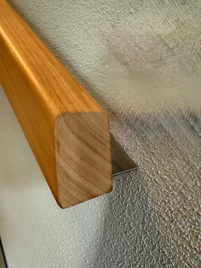 Nahaufnahme eines hölzernen Handlaufs, der mit einer Metallhalterung an einer strukturierten Wand befestigt ist. Der Handlauf hat eine glatte, rechteckige Form mit abgerundeten Kanten.