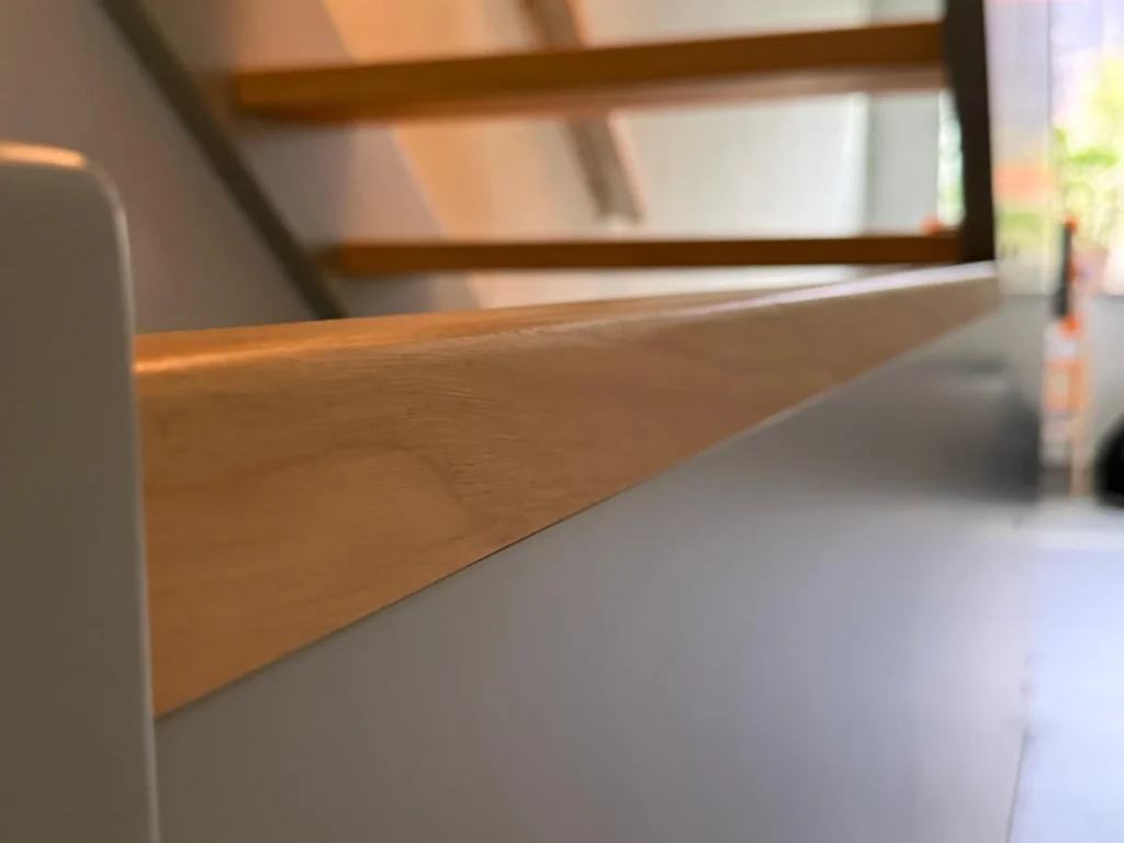 Nahaufnahme eines hölzernen Handlaufs auf einer Treppe, mit unscharfen Holzstufen und einem hellen Fenster im Hintergrund. Der Schwerpunkt liegt auf der glatten Textur des Handlaufs.
