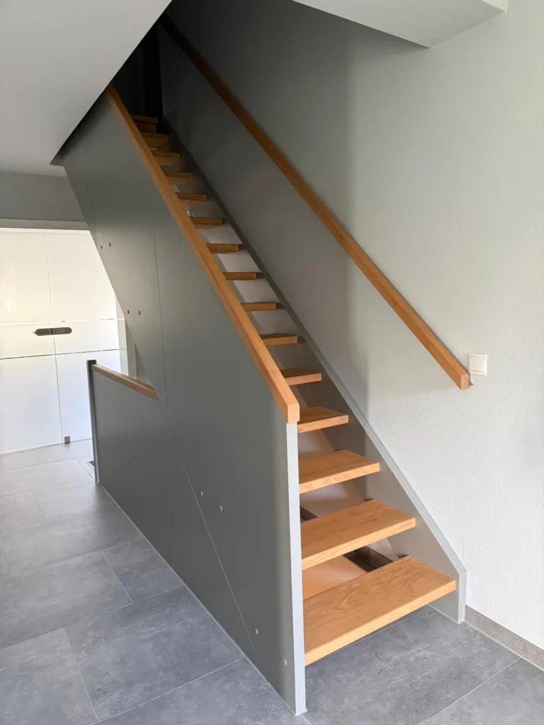 Eine moderne Innentreppe mit hellen Holzstufen und Handläufen, grauen Seitenwänden und hellgrauen Wänden, die von einem gefliesten Boden in einem sauberen, minimalistischen Innenraum nach oben führt.