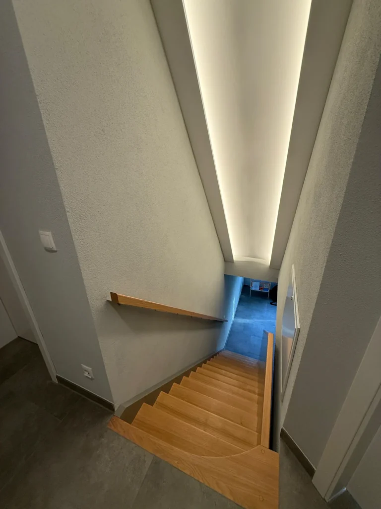Eine moderne Innentreppe mit Holzstufen, hellen Wänden und einem Holzhandlauf auf der linken Seite. An der Decke über der Treppe ist eine lange, helle Einbauleuchte angebracht.