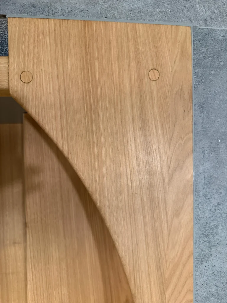 Nahaufnahme einer Holzoberfläche mit sichtbarer Maserung, zwei runden Holzdübeln und einem geschwungenen Detail auf der linken Seite, vor einem grauen Beton- oder Steinhintergrund.