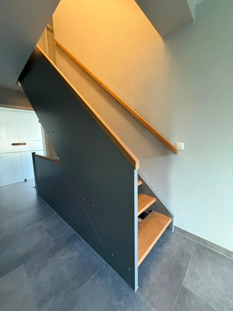 Moderne Innentreppe mit Holzstufen, einer schwarzen Seitenwand und einem Holzhandlauf vor hellen Wänden und grauem Fliesenboden. Der Raum ist durch natürliches und künstliches Licht hell erleuchtet.