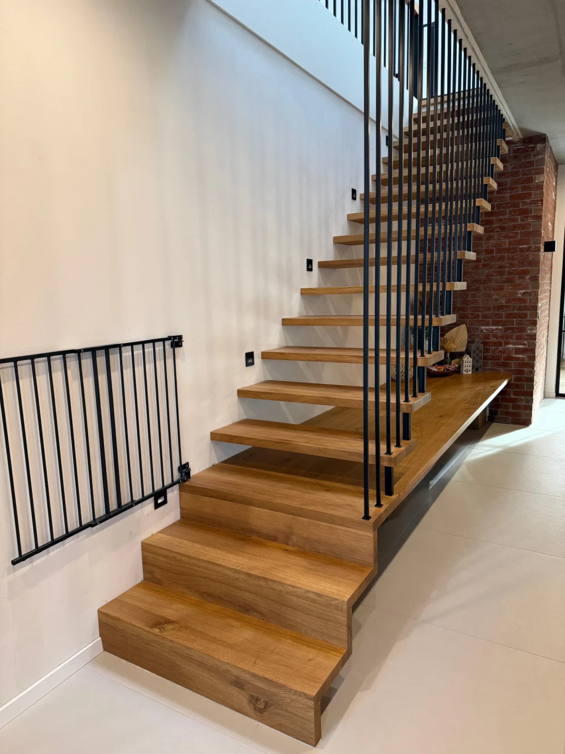 Eine moderne Holztreppe mit offenen Setzstufen und vertikalen schwarzen Metallgeländern führt neben einer weißen Wand und Sichtmauerwerk in einem zeitgenössischen Innenraum nach oben.