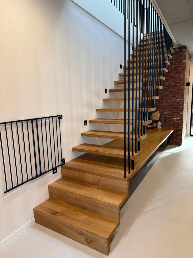 Eine moderne Holztreppe mit offenen Setzstufen und vertikalen schwarzen Metallgeländern führt neben einer weißen Wand und Sichtmauerwerk in einem zeitgenössischen Innenraum nach oben.