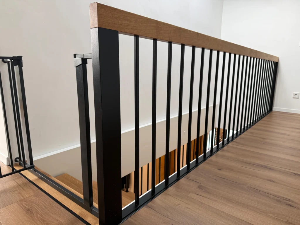 Ein modernes Treppengeländer mit schwarzen Metallstäben und einem hölzernen Handlauf, das auf einem Treppenabsatz mit Holzfußboden neben einer nach unten führenden Treppe angebracht ist. Am oberen Ende der Treppe ist ein Sicherheitsgitter angebracht.