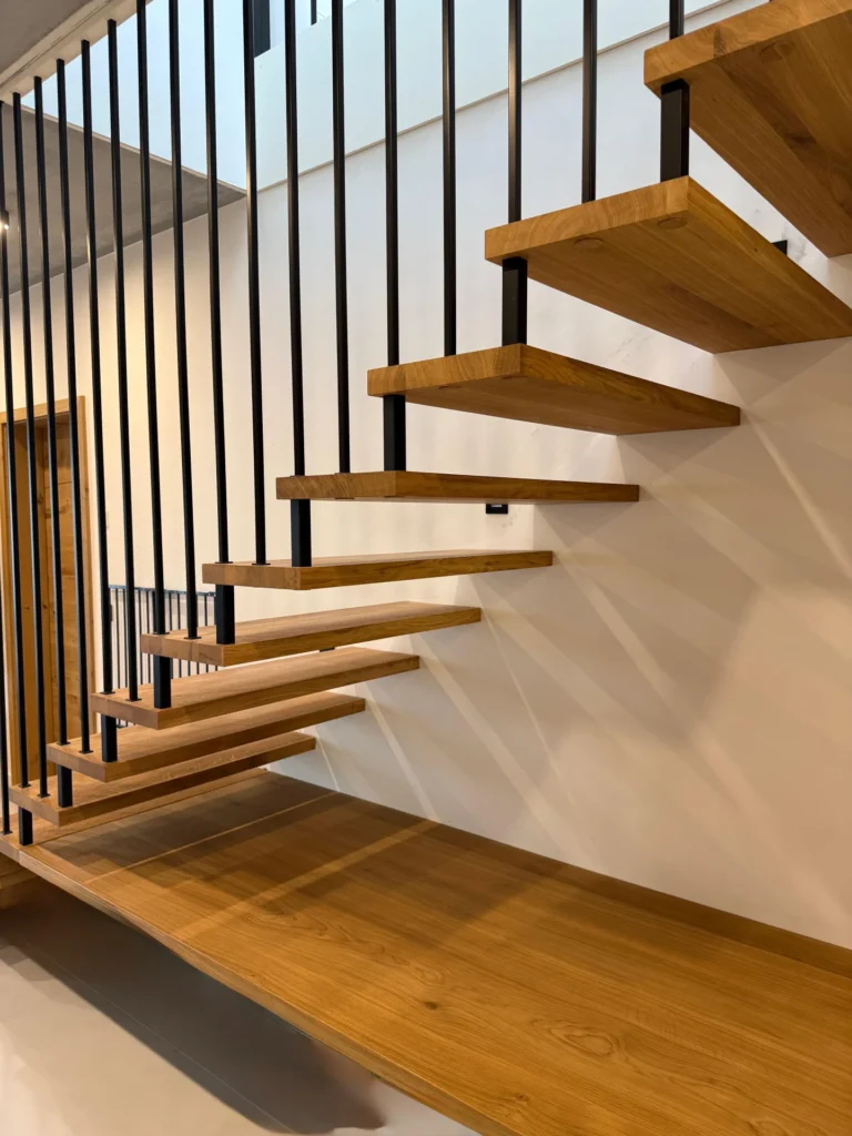 Eine moderne schwebende Holztreppe mit offenen Setzstufen und schwarzem vertikalen Metallgeländer, die an einer weißen Wand in einem zeitgenössischen Innenraum angebracht ist.