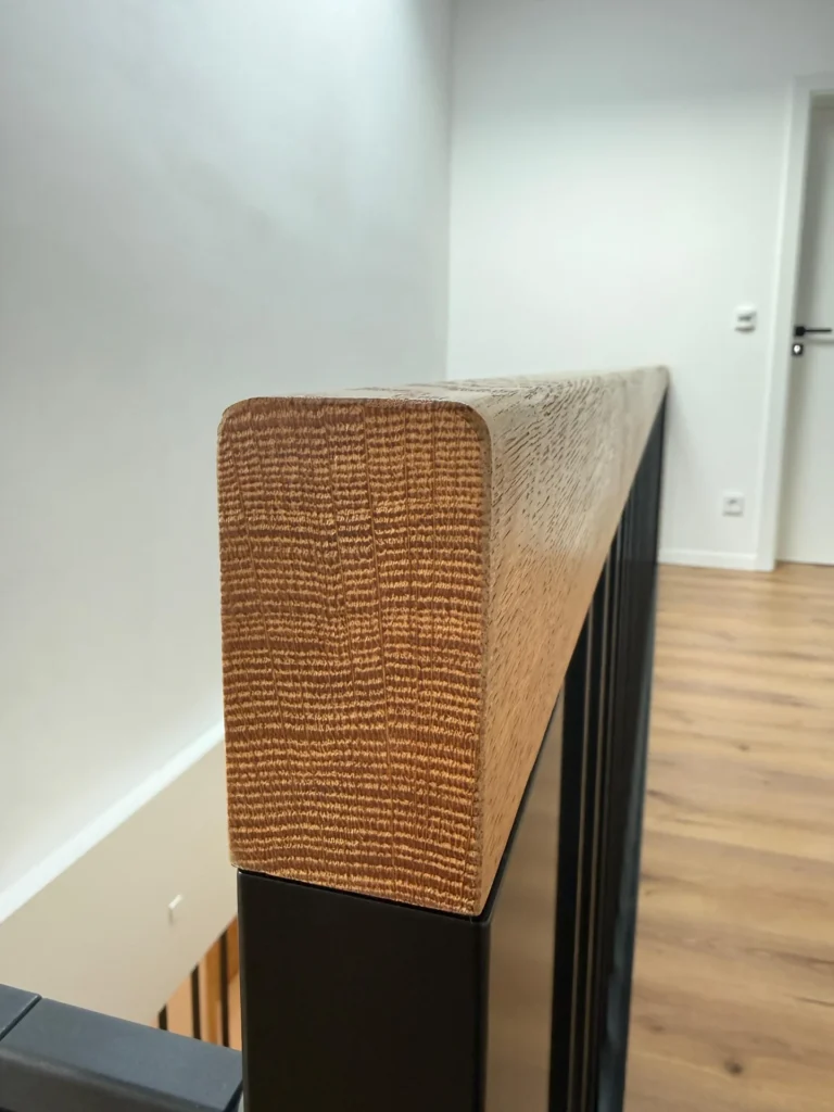Nahaufnahme eines hölzernen Handlaufs mit sichtbarer Maserung, montiert auf schwarzen Metallbalustern, in einem modernen Interieur mit weißen Wänden und hellem Holzfußboden. Das Foto konzentriert sich auf das Ende des Handlaufs.