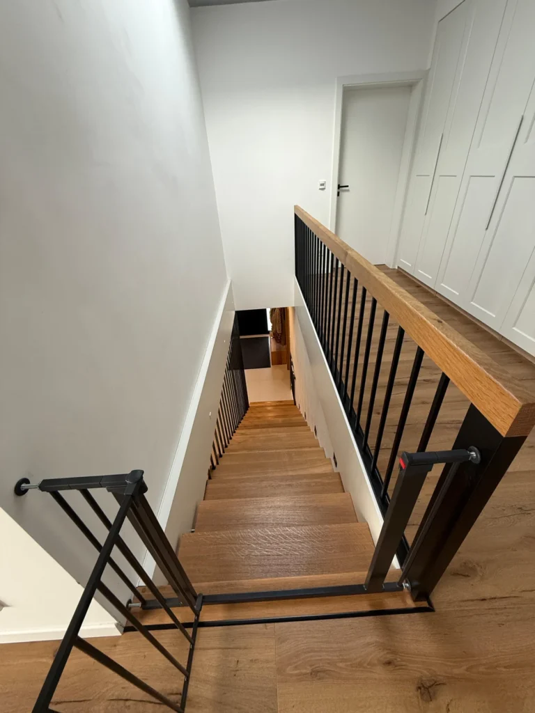 Blick von oben auf eine Treppe mit Holzstufen und schwarzem Geländer, die oben mit einem Sicherheitsgitter versehen ist. Die Treppe führt hinunter in einen Flur mit weißen Wänden und Holzfußboden. Im Obergeschoss ist eine geschlossene weiße Tür zu sehen.