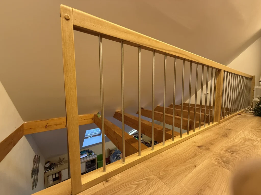 Holzgeländer mit vertikalen Metallstäben in einem oberen Stockwerk, mit Blick auf einen darunter liegenden Raum mit sichtbaren Deckenbalken, Holzboden und Regalen im Hintergrund.