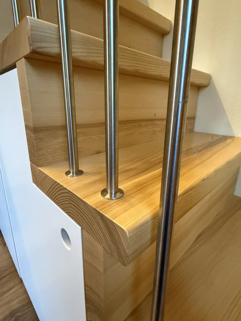 Nahaufnahme einer Holztreppe mit hellen Stufen und vertikalen Metallgeländern. Das moderne Design zeichnet sich durch klare Linien, eine weiße Verkleidung und einen runden Ausschnitt an der Seite aus.