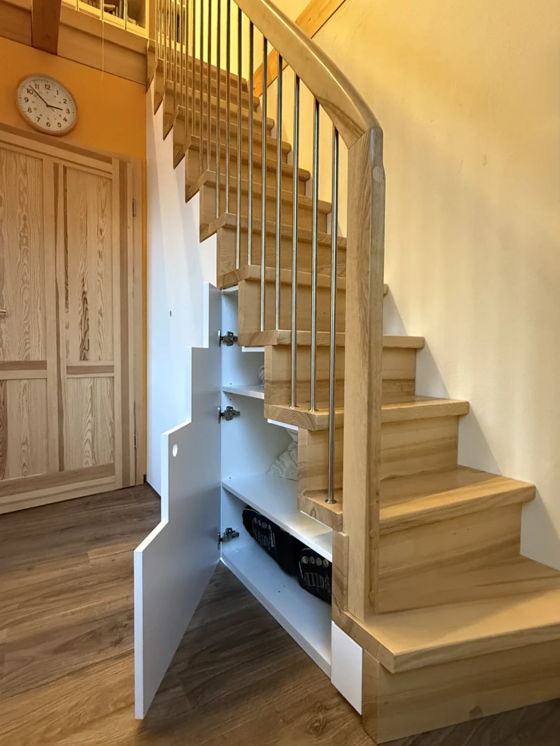 Eine Holztreppe mit einem eingebauten Schrank unter den Stufen, mit weißen Regalen und einer offenen Tür; im Hintergrund sind eine Uhr und geschlossene Flügeltüren zu sehen.