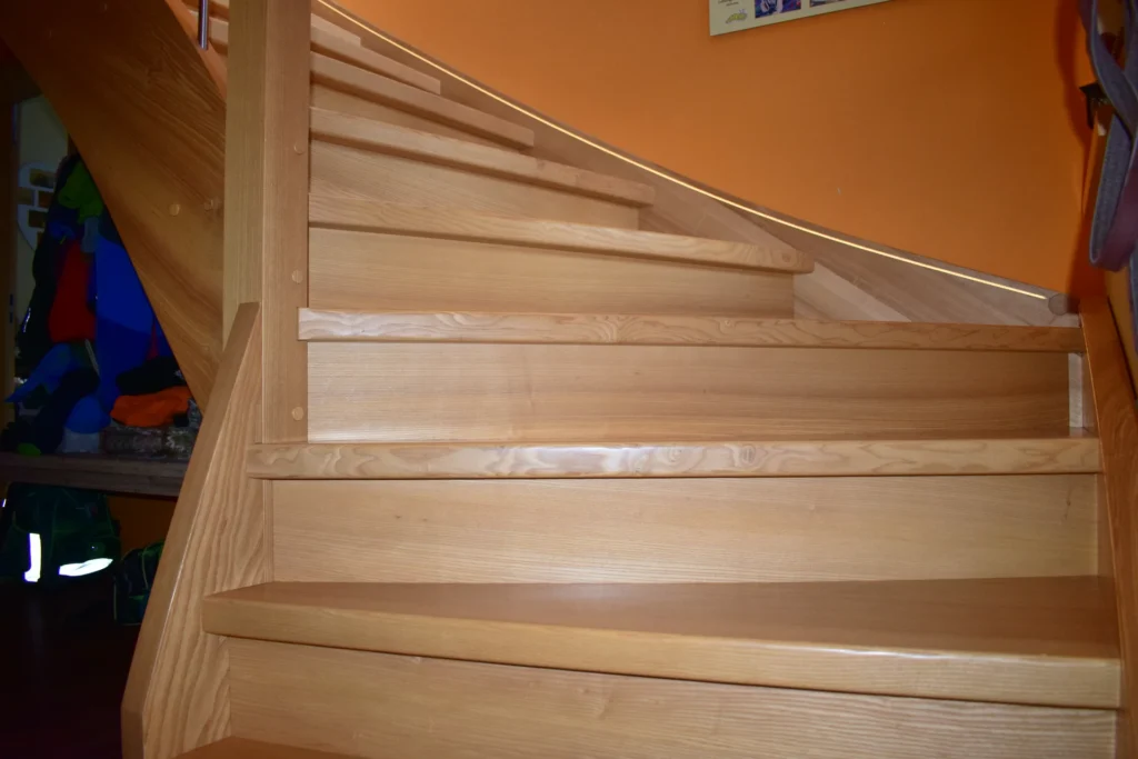 Nahaufnahme einer Holztreppe mit hellen Stufen und einer orangefarbenen Wand im Hintergrund. Die Treppe wölbt sich nach links aufwärts, und auf der linken Seite ist ein Teil eines Handlaufs zu sehen.