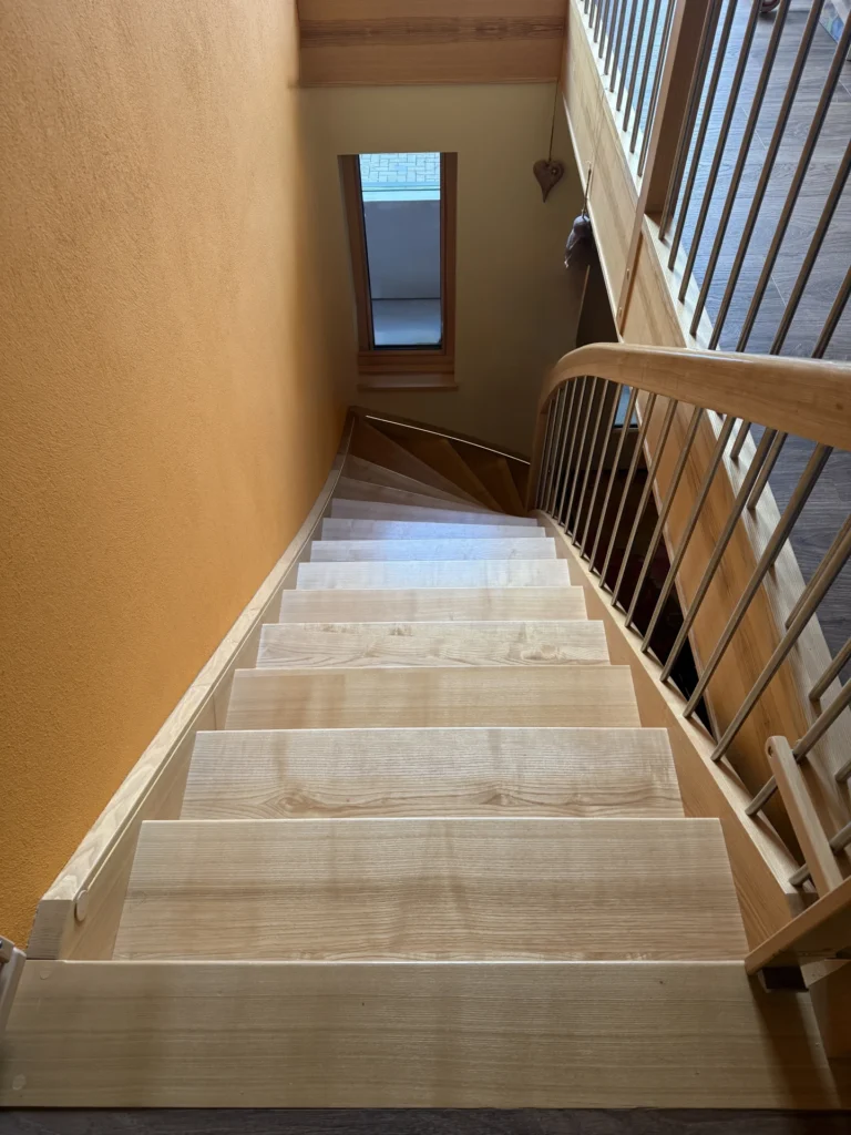Blick auf eine Holztreppe mit geschwungenem Geländer, gelb-orangefarbener Wand auf der linken Seite und einem Fenster am unteren Ende der Treppe, das Tageslicht hereinlässt.