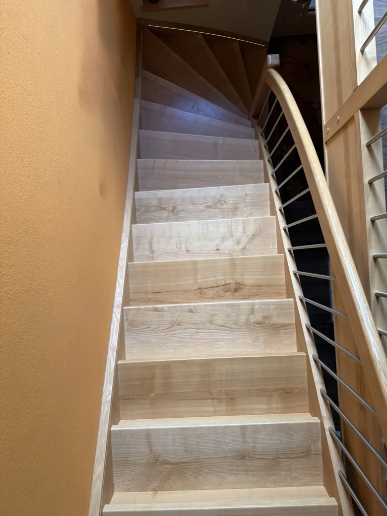 Eine moderne Holztreppe mit hellen Stufen, einem geschwungenen Geländer auf der rechten Seite und einer leuchtend orangefarbenen Wand auf der linken Seite, von unten nach oben gesehen. Durch ein Fenster auf der rechten Seite fällt natürliches Licht ein.
