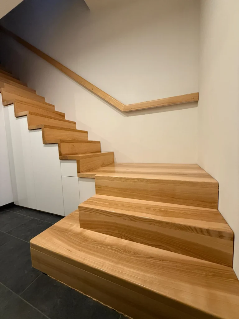 Eine moderne Treppe mit breiten Holzstufen und einem minimalistischen Holzhandlauf vor einer weißen Wand. Unter der Treppe befinden sich weiße Einbauschränke und ein dunkel gefliester Boden.