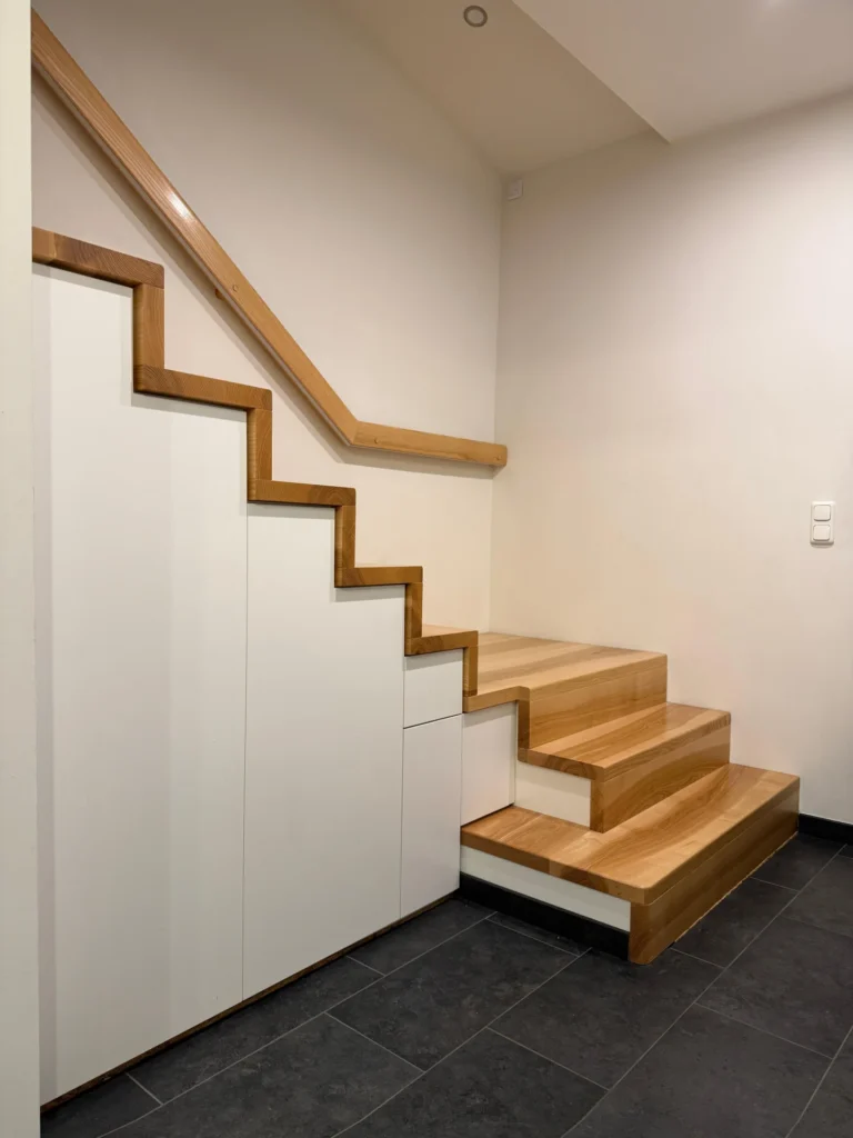 Holztreppe mit eingebauten weißen Schränken darunter, die ein modernes und funktionales Design schaffen. Die Stufen und der Handlauf sind aus hellem Holz, der Bodenbelag besteht aus dunklen Kacheln. Die Wände sind schlicht und hell.
