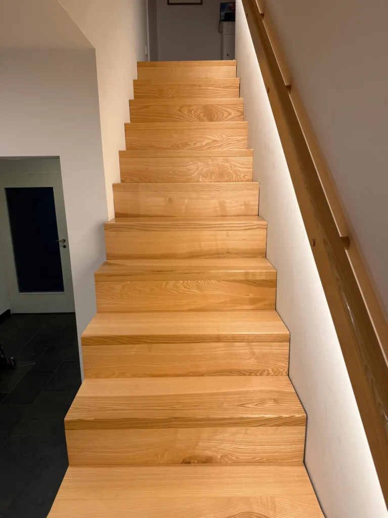 Eine gerade Treppe aus hellem Holz mit einem passenden Holzhandlauf auf der rechten Seite, die zu einem hellen oberen Flur hinaufführt. Die Wände sind weiß und der Raum wirkt modern und sauber.