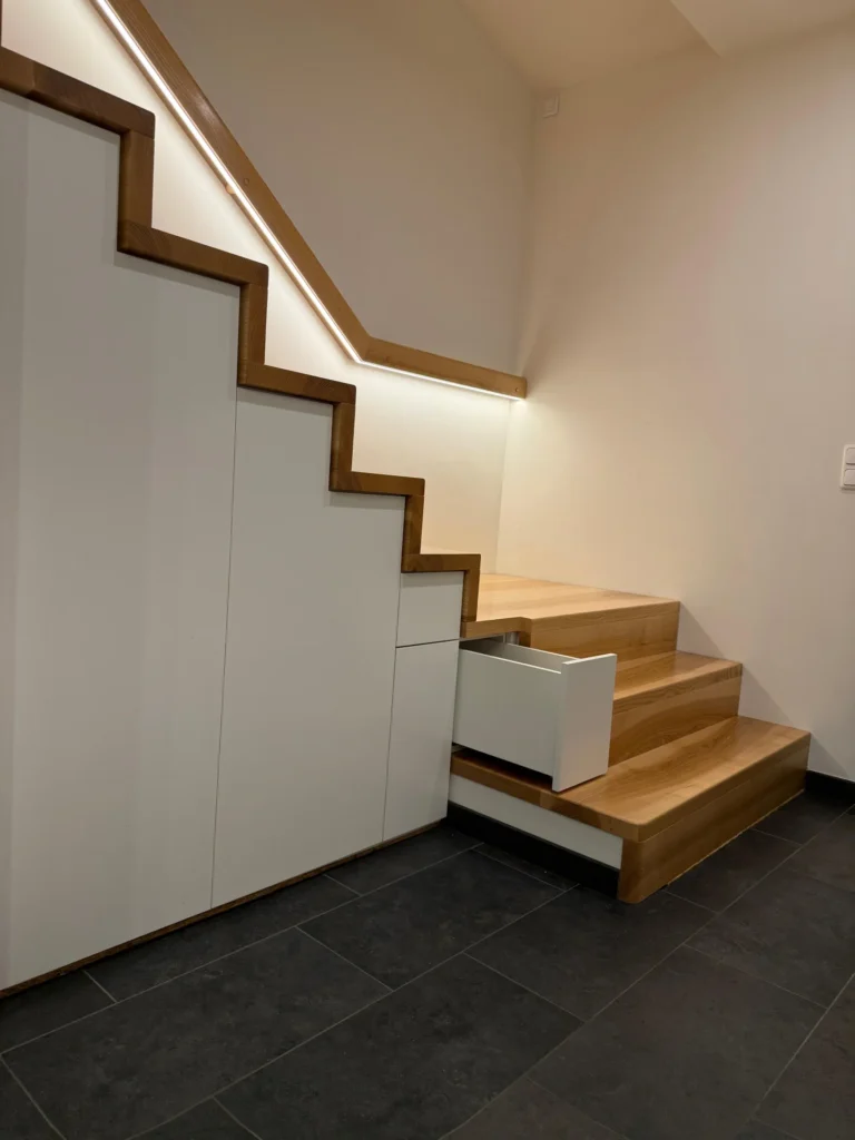 Eine moderne Holztreppe mit eingebauten weißen Schränken, darunter eine offene Schublade; warme Beleuchtung an der Seite der Treppe und dunkle Fliesen auf dem Boden.
