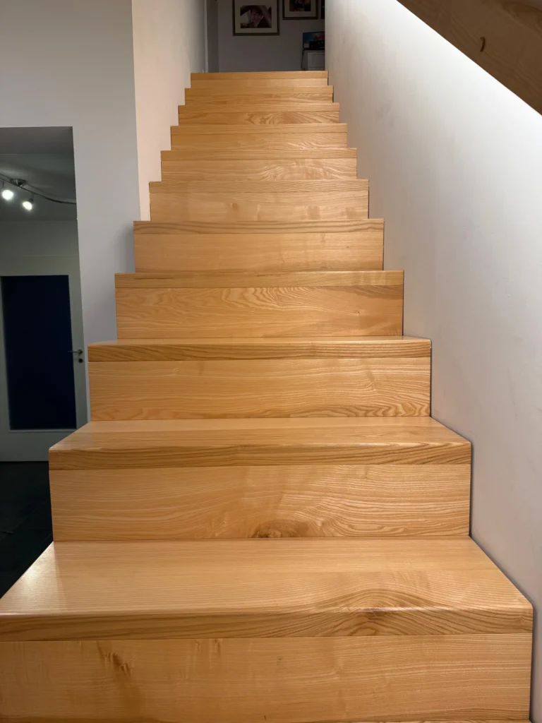 Eine Treppe aus hellem Holz führt in einem modernen Interieur zwischen weißen Wänden nach oben. Die Stufen sind breit und glatt, und an der oberen Wand sind gerahmte Bilder zu sehen.