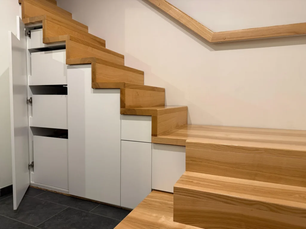Eine moderne Holztreppe mit eingebauten weißen Schränken und Schubladen darunter, von denen einige offen sind, was eine effiziente Raumausnutzung zeigt.