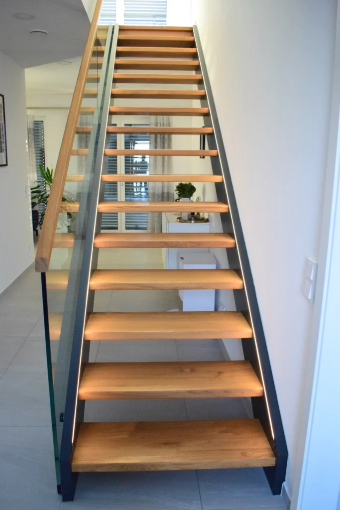 Eine moderne Holztreppe mit offenen Setzstufen und Glasgeländer, die von dezenten LED-Lichtbändern beleuchtet wird, führt zu einem oberen Stockwerk in einem hellen, zeitgenössischen Innenraum.