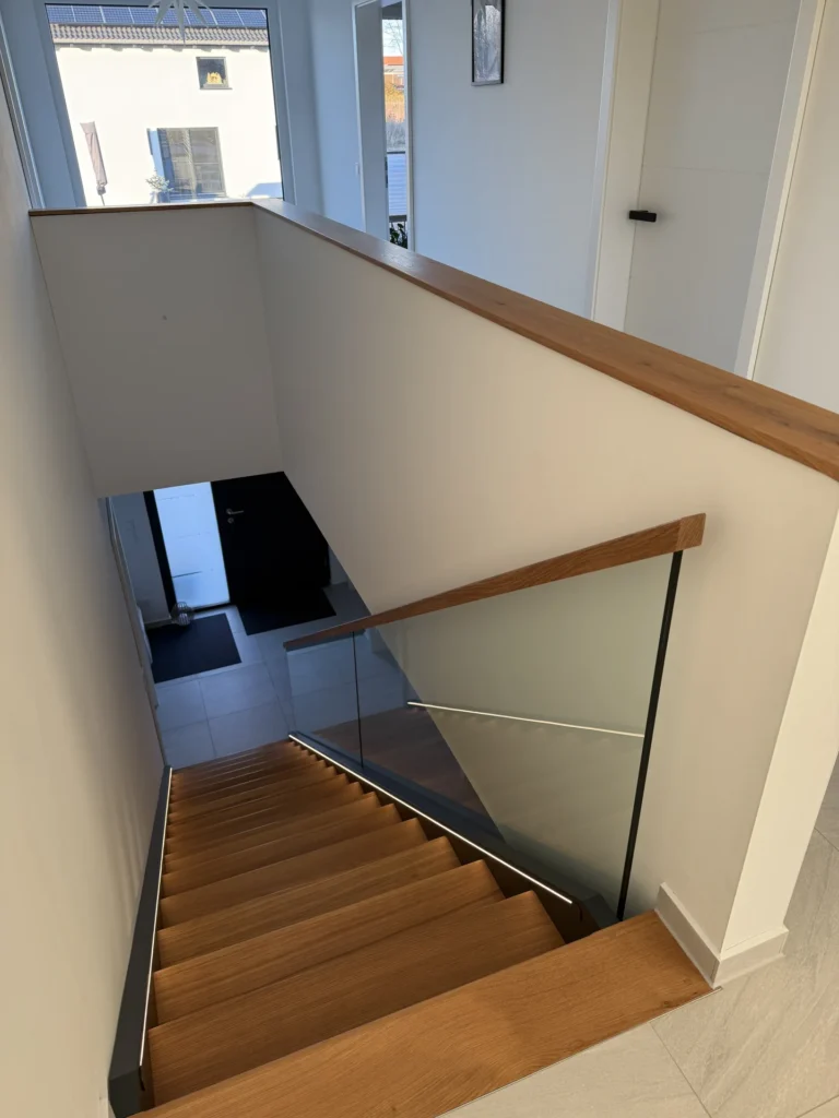 Eine moderne Innentreppe mit Holzstufen und Glasgeländer, die zu einem gefliesten Eingangsbereich mit schwarzer Tür und natürlichem Licht aus den Fenstern führt.