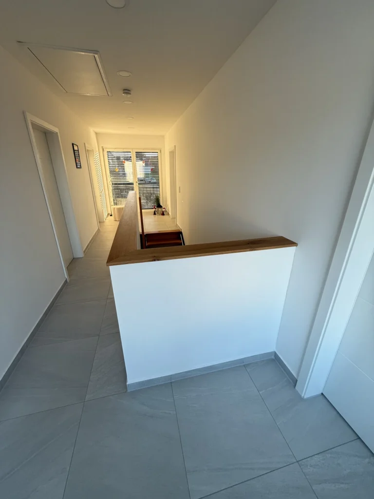 Ein heller Flur mit hellgrauen Fliesenböden, weißen Wänden und einer hölzernen Arbeitsplatte entlang einer halben Wand. Am Ende führen Glastüren zu einem Balkon, auf den das Sonnenlicht fällt. Auf beiden Seiten des Flurs befinden sich Türen.