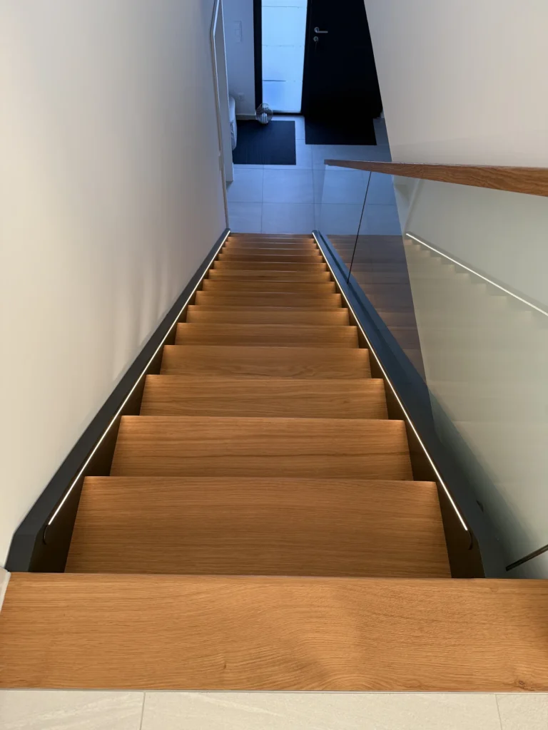 Blick auf eine moderne Holztreppe mit eingebauten LED-Lichtbändern an den Seiten, einem Glasgeländer auf der rechten Seite und einer schwarzen Tür am unteren Ende der Treppe.