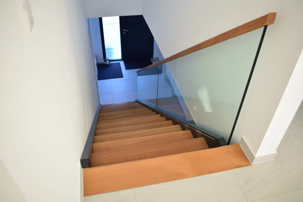 Eine Holztreppe mit Glasgeländer führt hinunter zu einem gefliesten Eingang mit geschlossener schwarzer Tür; Sonnenlicht beleuchtet das moderne, minimalistische Interieur.