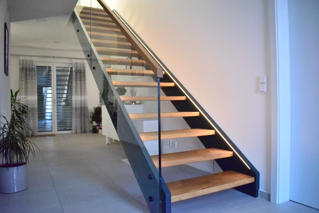 Eine moderne Innentreppe mit Holzstufen, Glaswänden und LED-Lichtbändern, die in einem hellen, minimalistischen Flur mit Pflanzen und großen Fenstern im Hintergrund zu einem oberen Stockwerk führt.