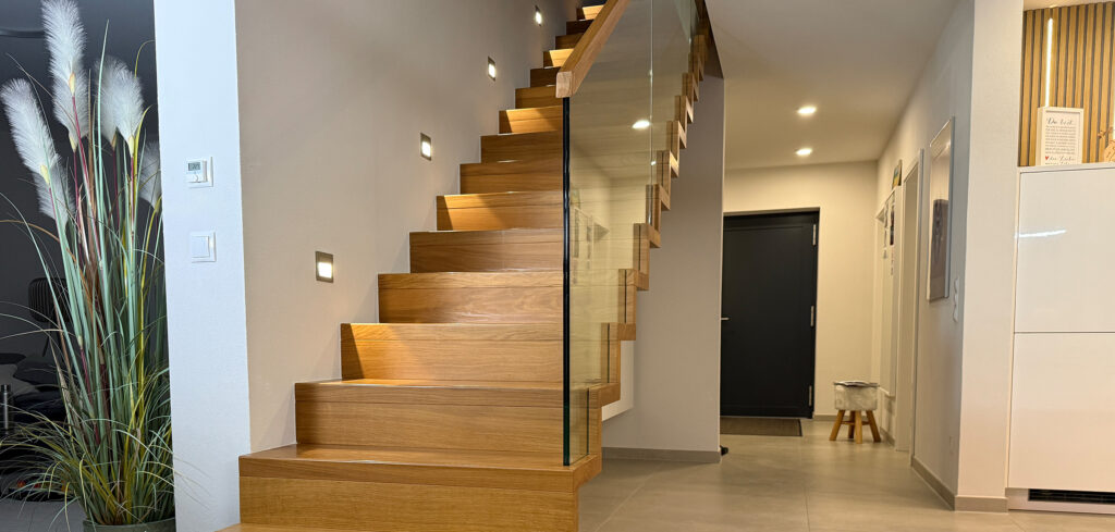 Moderne Holztreppe mit Glasgeländer, montiert an einer weißen Wand mit eingebauten Stufenleuchten. Links steht hohes Ziergras in einer Vase; im Hintergrund sind ein Flur und eine schwarze Tür zu sehen.