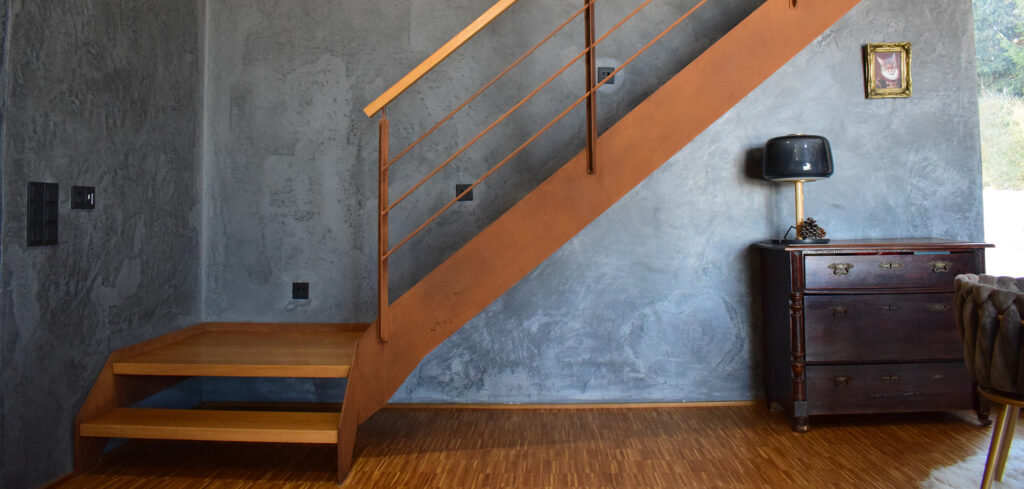 Ein modernes Interieur mit einer Holztreppe, einer grau strukturierten Wand, einer kleinen antiken Kommode mit einer Lampe und einem gerahmten Bild über der Kommode. Von der rechten Seite fällt Licht ein.