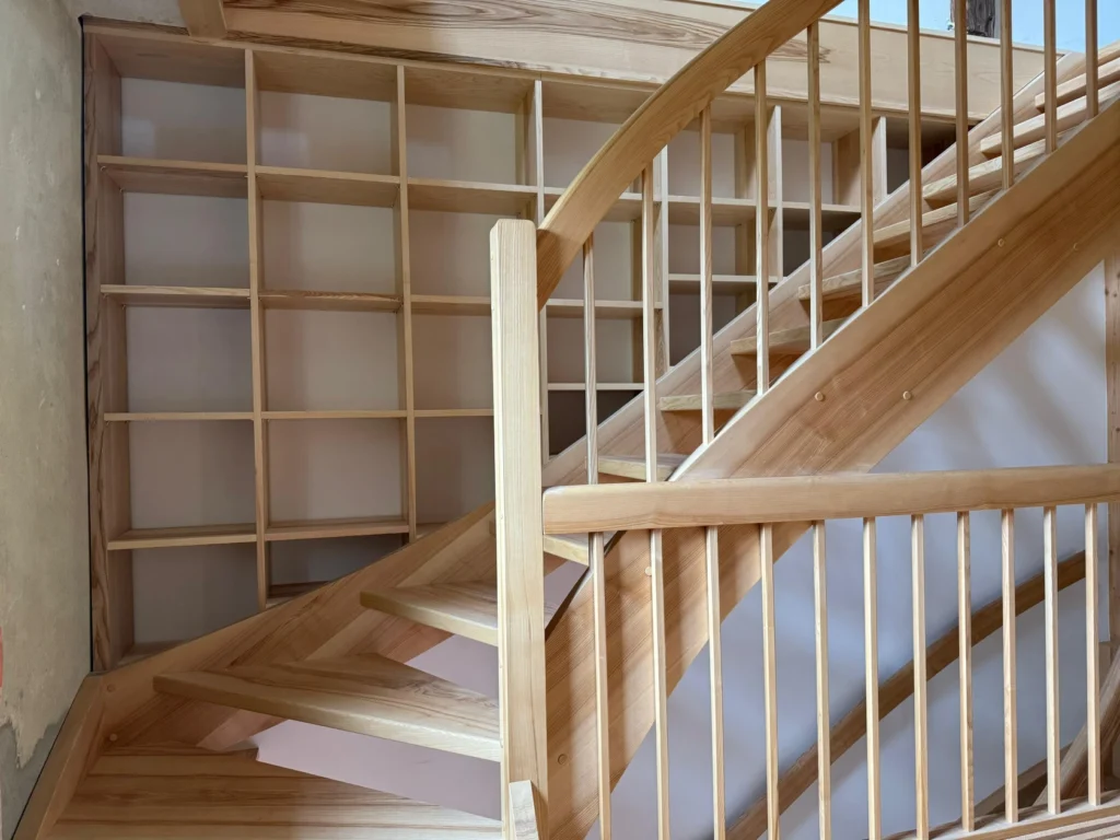 Eine Holztreppe mit vertikalen und horizontalen Latten und eingebauten Ablagefächern unter der Treppe. Das Design betont natürliche Holztöne und klare, geometrische Linien.