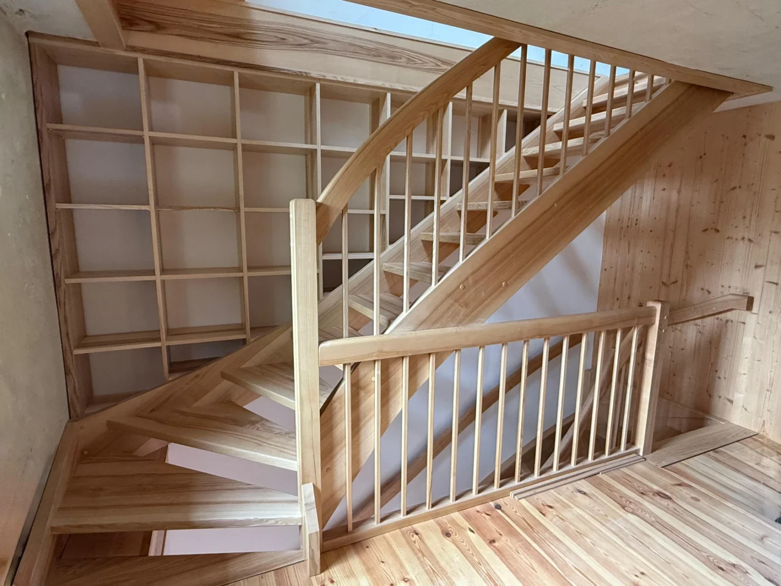 Eine Holztreppe aus hellem Holz mit schrägen Stufen, senkrechten Geländern und einer geometrischen Gitterwand daneben, alles in einem warm beleuchteten Innenraum.