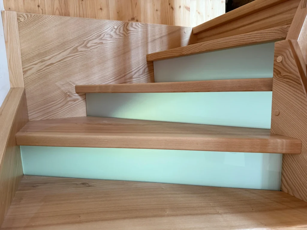 Nahaufnahme einer modernen Holztreppe mit hellen Holzstufen und weißen vertikalen Setzstufen vor einer holzgetäfelten Wand.