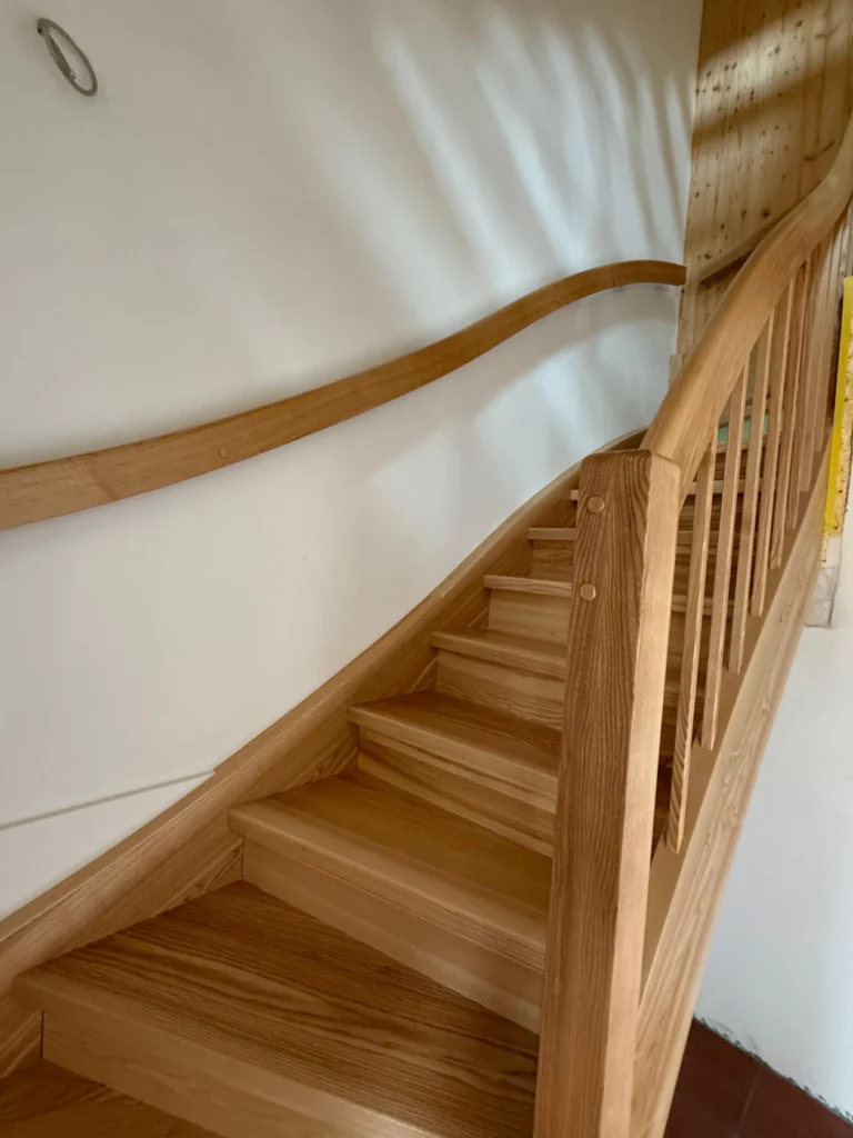 Eine Holztreppe mit geschwungenem Handlauf und vertikalen Geländerstäben führt entlang einer weißen Wand in ein höheres Stockwerk. Das Licht erzeugt weiche Schatten an der Wand.