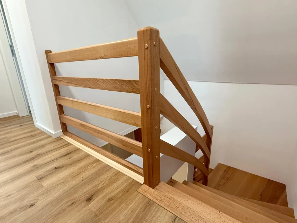 Eine Holztreppe mit horizontalem Geländer und Naturholzboden, die von einem oberen Treppenabsatz hinunterführt. Die Wände sind weiß, was ein helles und sauberes Erscheinungsbild schafft.