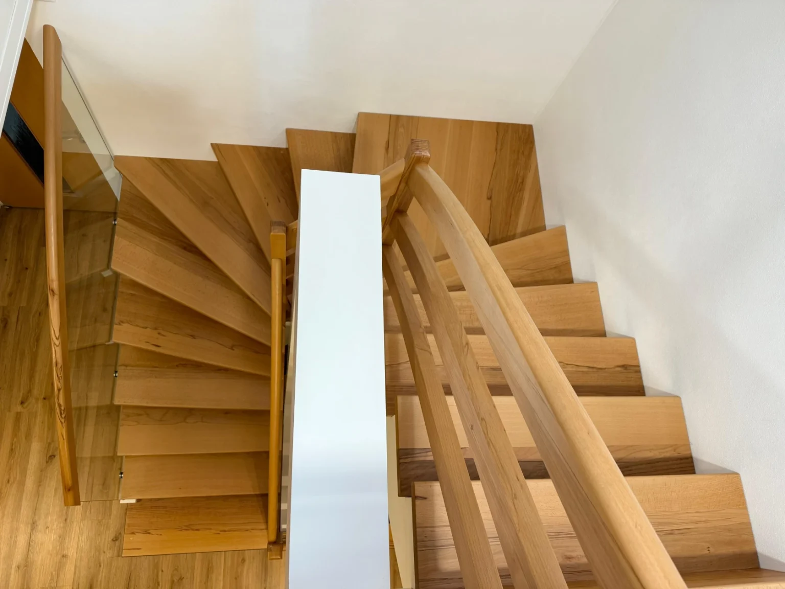 Eine Holztreppe mit einem geschwungenen Handlauf, von oben gesehen. Die Stufen drehen sich im rechten Winkel um eine weiße Mittelstütze, wobei der Holzboden am oberen und unteren Ende der Treppe sichtbar ist.