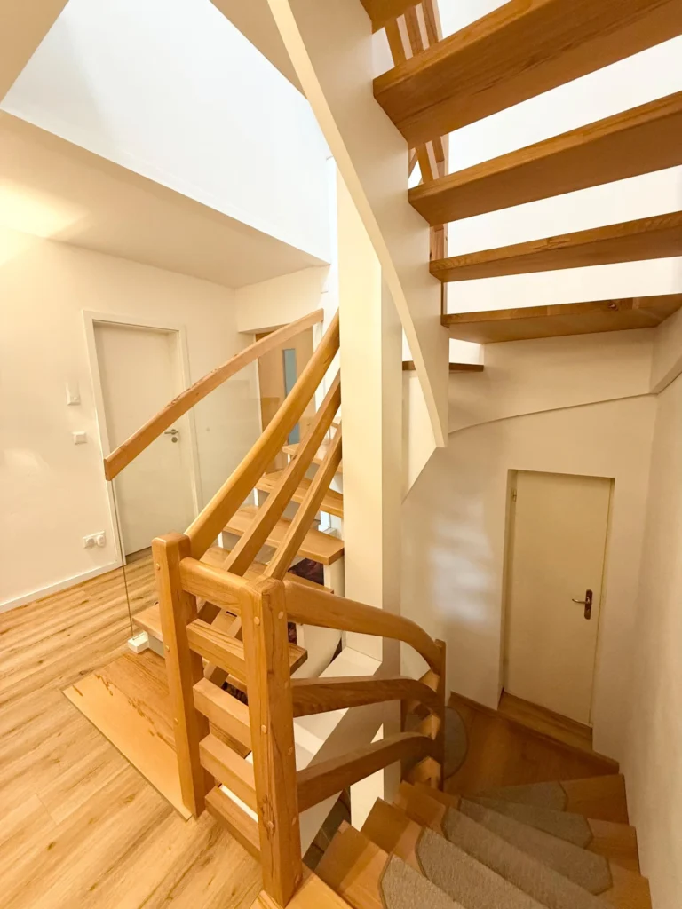 Eine moderne Innentreppe aus Holz mit hellbraunen Stufen und Handläufen, die sich durch einen hellen Innenraum mit weißen Wänden, Holzböden und einer kleinen Tür unter der Treppe nach oben windet.
