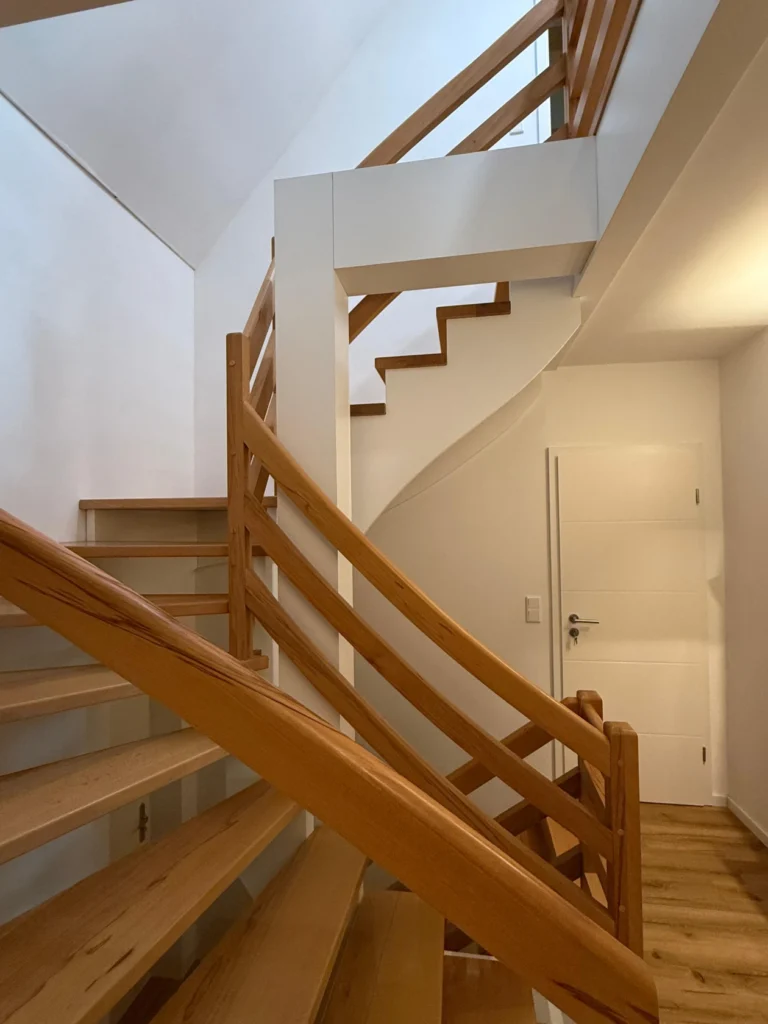 Eine moderne Holztreppe mit hellbraunen Handläufen und Stufen schlängelt sich in einem weiß getünchten Innenraum nach oben, wobei auf der rechten Seite eine geschlossene weiße Tür zu sehen ist.