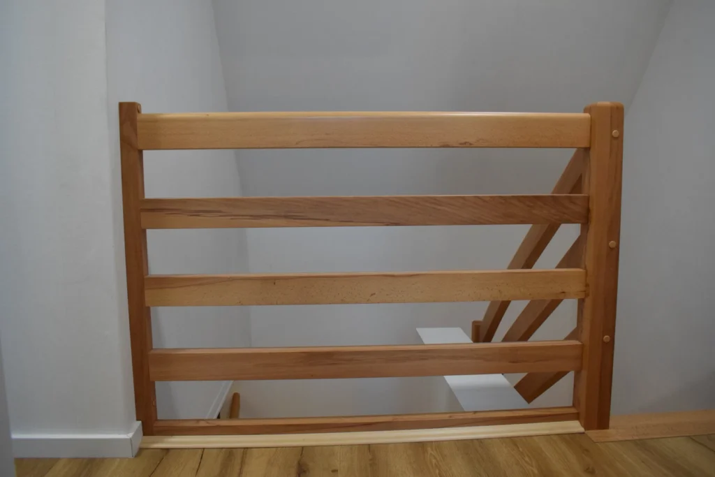 Ein hölzernes Sicherheitstor mit horizontalen Lamellen blockiert den oberen Teil einer Treppe. Das Tor ist auf einem hellen Holzboden neben einer weißen Wand installiert und verhindert den Zugang zur darunter liegenden Treppe.