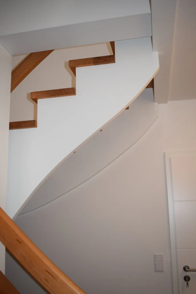 Modern gestaltete Holztreppe mit weißer Unterseite und hellen Holzstufen und Geländern, die sich entlang einer weißen Wand in einem Wohnbereich nach oben schlängelt. Darunter sind eine weiße Tür und ein Lichtschalter zu sehen.
