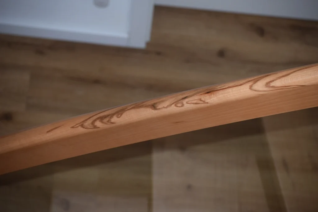 Nahaufnahme eines hölzernen Handlaufs mit sichtbaren Maserungen vor einem unscharfen Hintergrund eines hellen Holzbodens und einer weißen Wand.