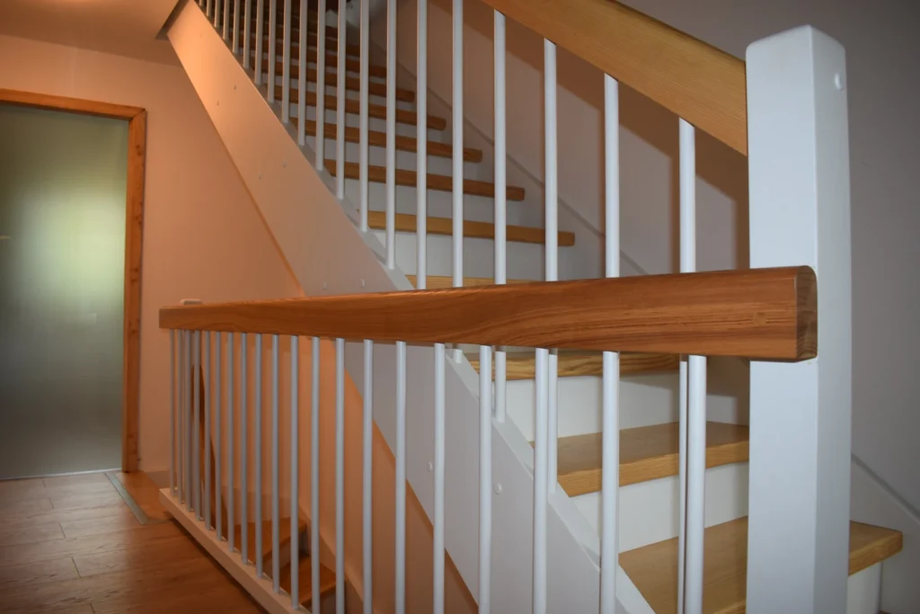 Eine moderne Innentreppe aus Holz mit weißem vertikalem Geländer und hellbraunen Stufen und Handlauf, die in einem hellen, minimalistischen Wohnbereich in die nächste Etage führt.
