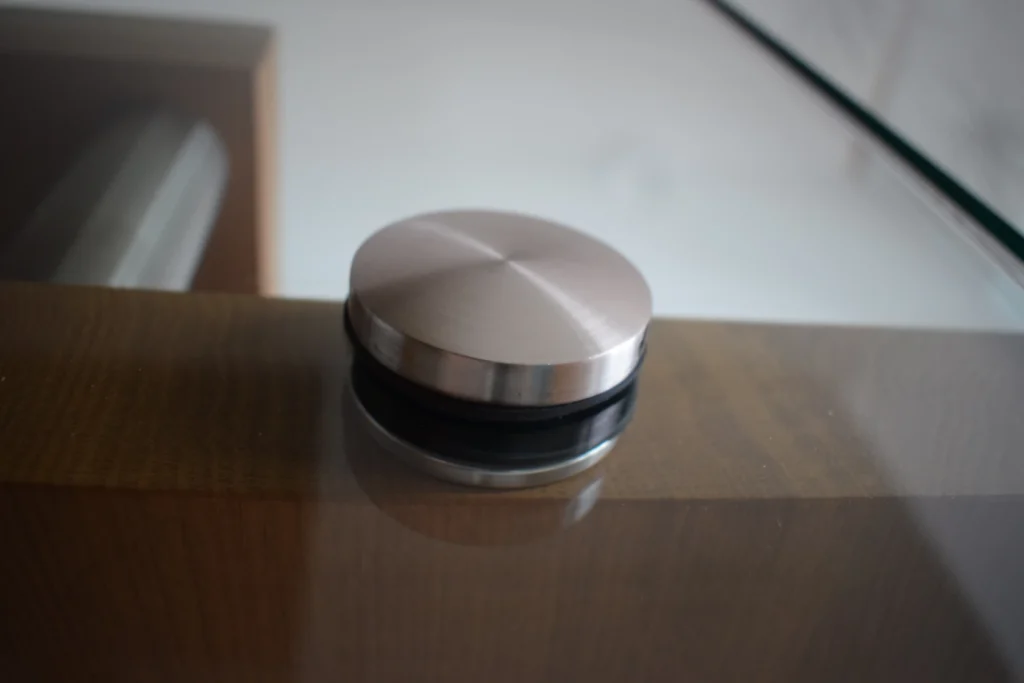 Nahaufnahme eines runden Metallbeschlags, mit dem eine Glasscheibe an einer Holzoberfläche befestigt ist, wahrscheinlich Teil eines Geländers oder einer Trennwand. Der Beschlag hat eine gebürstete Metalloberfläche und eine schwarze Gummidichtung.