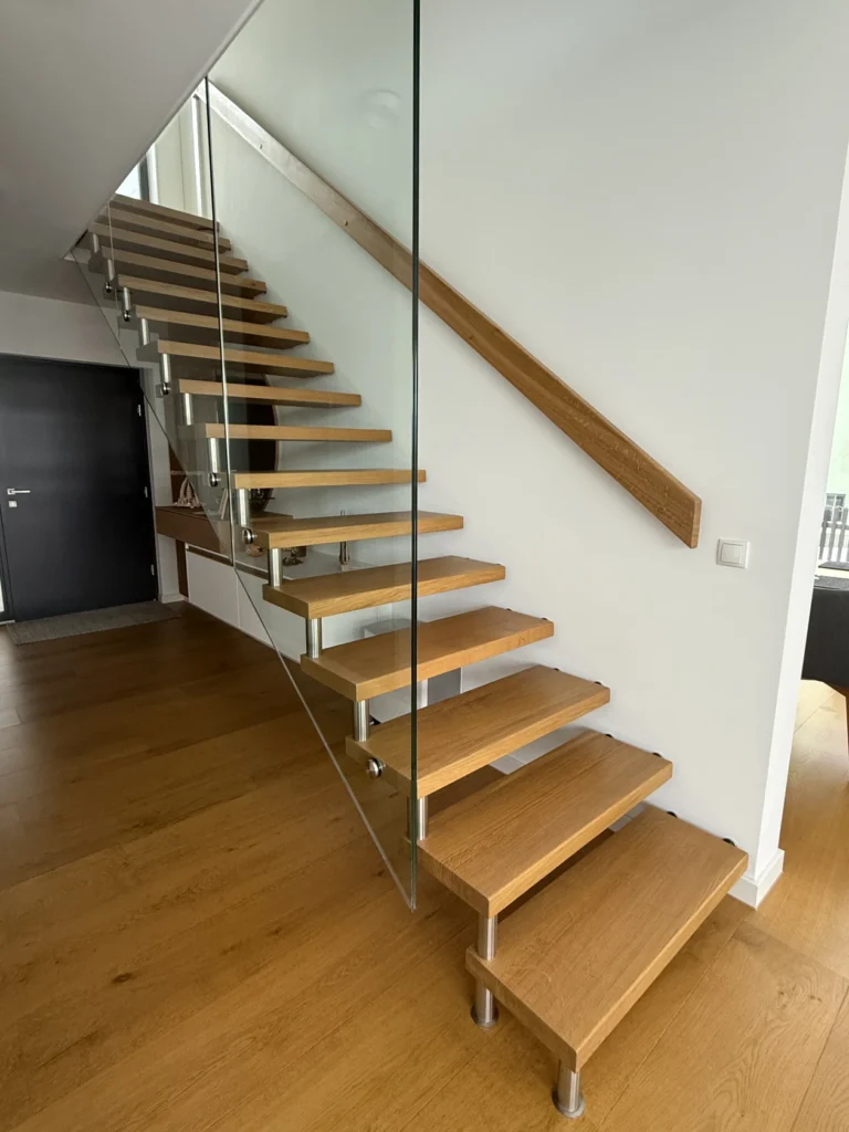 Eine moderne schwebende Treppe mit Holzstufen, Metallstützen und einer Seitenwand aus Glas vor weißen Wänden und einem Holzboden in einem modernen Haus.