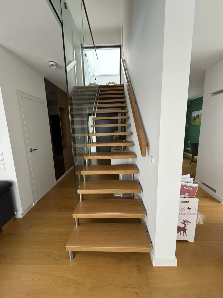 Eine moderne Innentreppe mit schwimmenden Holzstufen und einem Glasgeländer, die entlang einer weißen Wand in einer hellen, modernen Wohnung mit Holzfußboden hinaufführt. Rechts am unteren Ende der Treppe sind Kisten gestapelt.