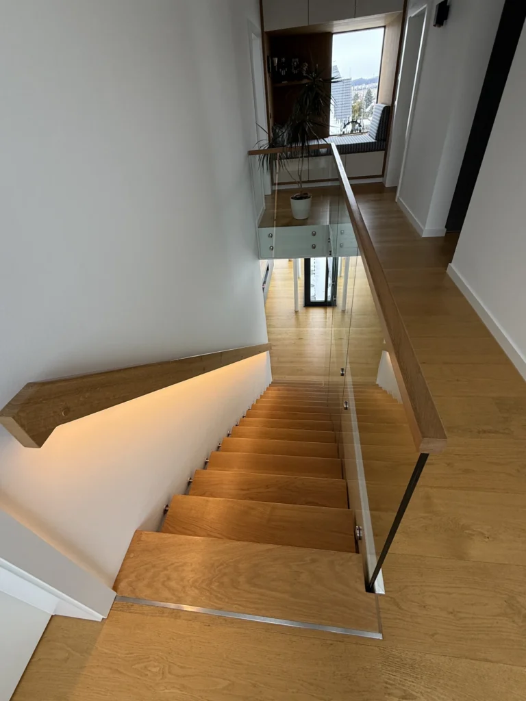 Eine moderne Innentreppe aus Holz mit Glasgeländer und integrierter Beleuchtung führt hinunter zu einem Flur mit Holzboden. Oben auf der Theke steht eine Topfpflanze, und im Hintergrund sind große Fenster zu sehen.