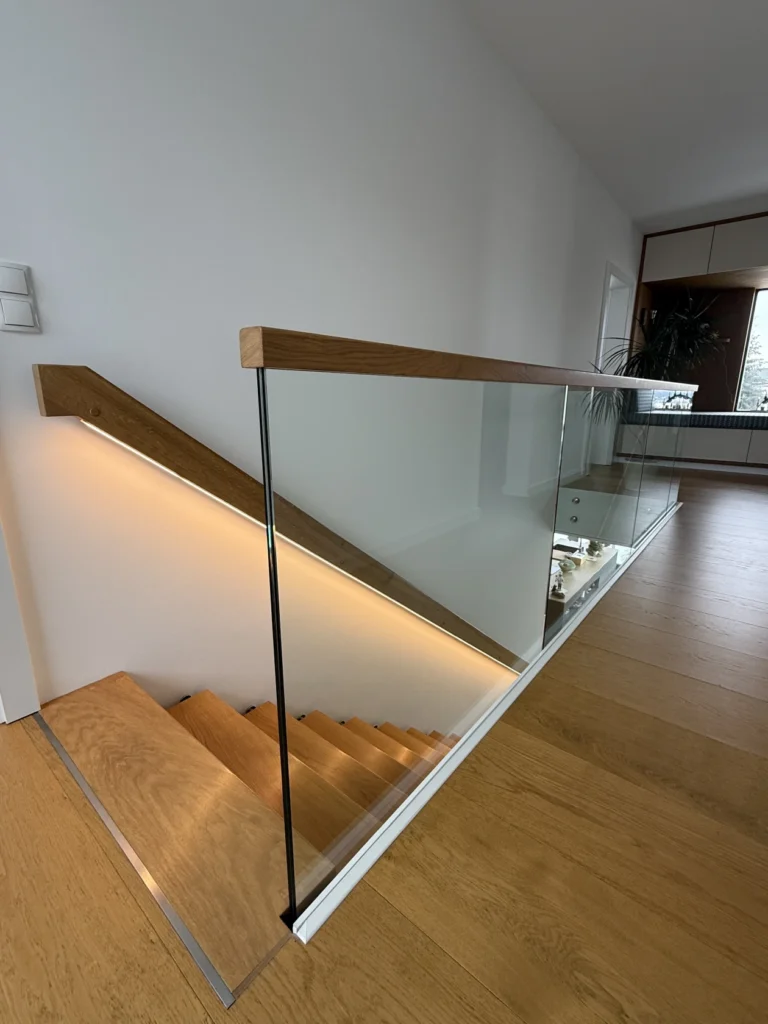 Moderne Holztreppe mit einem Glasgeländer und einem Holzhandlauf. Die Treppe verfügt über eine eingebaute Wandbeleuchtung, und der Flur hat einen hellen Holzboden und weiße Wände, die ein klares, minimalistisches Aussehen schaffen.